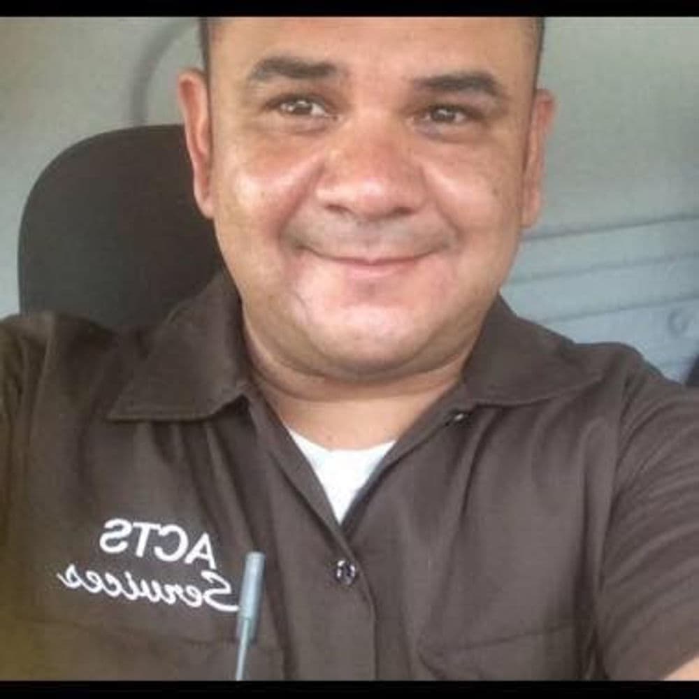 Male employee in a brown uniform shirt, smiling in a vehicle interior.