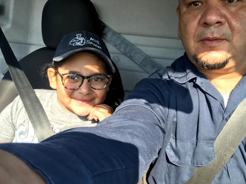 Girl and adult in vehicle, both wearing seatbelts, smiling at the camera.